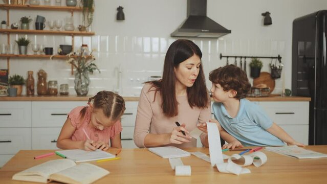 Single mother manages her finances by reviewing receipts, expenses and checks, trying to give her son attention and overseeing daughter's homework in kitchen. Challenging balance of single parenthood