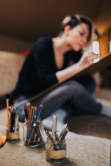 Close up of pencils in jars on sofa with artist in a background.
