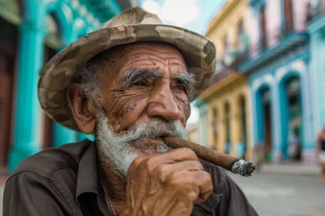 Foto op Plexiglas man in hat and glasses is smoking a cigar. Concept of relaxation and leisure, as the man is enjoying his cigarette while sitting on a chair. cuban cigar smoking old man, background Havana city houses © Nataliia_Trushchenko