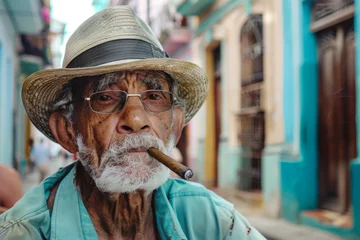 Poster man in hat and glasses is smoking a cigar. Concept of relaxation and leisure, as the man is enjoying his cigarette while sitting on a chair. cuban cigar smoking old man, background Havana city houses © Nataliia_Trushchenko