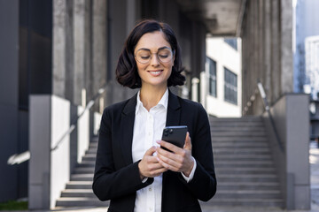 Fototapeta premium Close-up photo of a smiling young business woman in a suit and glasses standing outside an office building and using a mobile phone