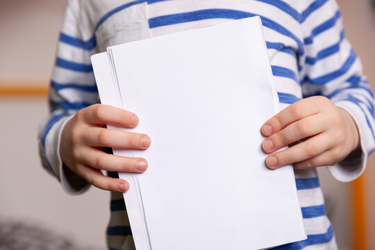 A closeup kid boy in a striped jacket holds white sheets of paper in his hands.