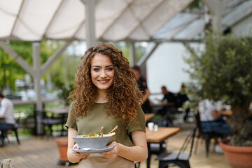 Portrait of a beautiful waitress holding plate with food, bowl with salad. Server standing on restaurant terrace in an apron. - 755175228