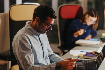 Business male person using mobile phone intently at desk in office setting, with female colleague working in the background.