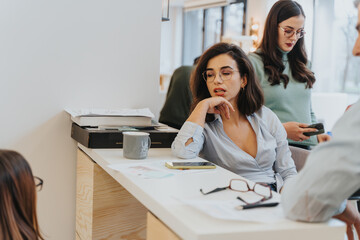 Pensive female professional in glasses at her desk with coworkers around in a contemporary...