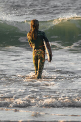 Young surfer girl in camo wetsuit entering the sea holding her surfboard