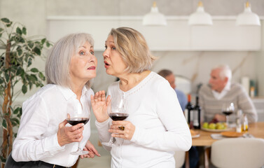 Two elderly women friends chatting and drinking wine in kitchen at home
