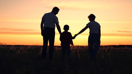 Happy family mom dad child walking in field on summer evening. Small kid holds his parents hands. Dad hand shows his son sunset. Parents hold their childs hands in outdoors. Concept family holidays