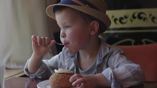 Seated at cafe table young boy in hat eating sweet cake. Sweet cake delight boy eating sweetness dances on his taste buds Boy eating sweet cake pastry completely immersed in joy of indulgence
