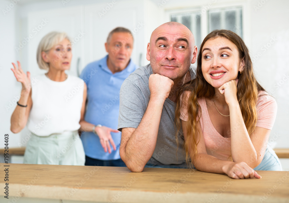 Wall mural Portrait of a happy married couple in a home kitchen, not paying attention to adult family members who reprimand them