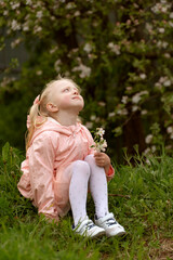 Portrait of little girl sitting on the ground and looking up at the sky. Child in blooming garden. Vertical frame.