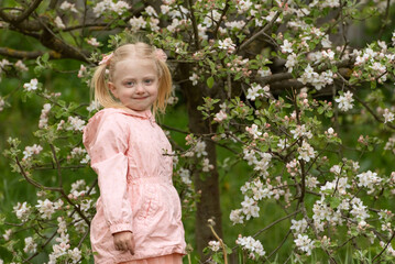 Portrait of cute beautiful blonde girl with two ponytails wears pink jacket stands in spring garden near flowering tree.