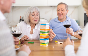 Fototapeta na wymiar Elderly happy people playing Jenga at home, woman is removing a wooden block from the tower and the others are watching