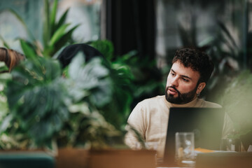 A concentrated man works on his laptop during a business team meeting at a cozy, plant-filled cafe....