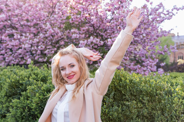 Young beautiful blond woman with standing in blooming sakura park raised her hands. Portrait of gentle girl in blossoming park.