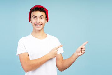 Attractive, happy teenage boy with braces wearing stylish red hat showing finger on copy space