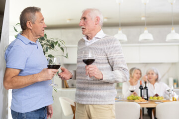 Fototapeta na wymiar During friendly meeting, two elderly men with glass of wine in their hands stand at distance from table, communicate and chatting, discussing sports news and hobby