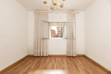 An empty room with light wooden floors with an open aluminum window and long dirty white curtains