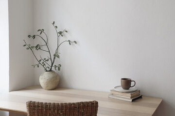 Scandinavian living room interior. Cup of coffee, books on wooden table. Willow tree branches, catkins in vase. Rattan chair. Elegant Scandi working space, home office. Blank beige wall background