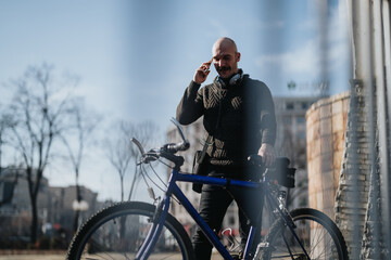 Stylish adult man in casual sweater making a phone call with his smart phone beside a blue bicycle...