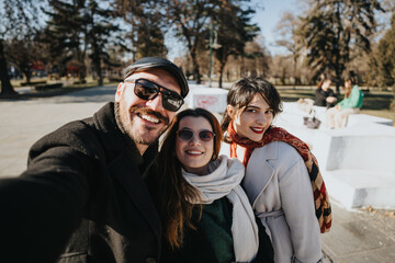 Three happy friends of diverse ethnicities sharing a joyful moment taking a group selfie on a sunny winter day in a public park.