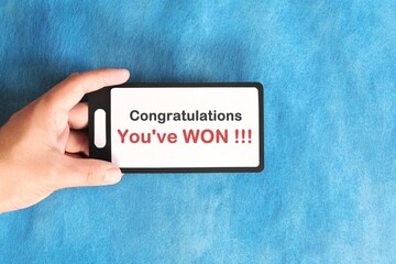 Winning text scam concept. Hand holding mobile phone with congratulatory message in blue background. - obrazy, fototapety, plakaty