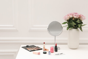 Mirror, cosmetic products and vase with pink roses on white dressing table in makeup room, space for text