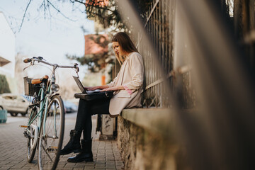 A young female professional working on a laptop seated outdoors on a sunny day with her bicycle...