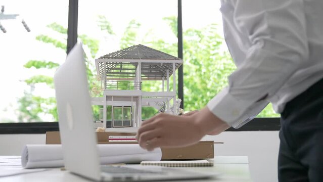 Close up of professional male architect engineer measure house model and using laptop analysis data at meeting table with house model and map . scatter around. Focus on hand. Immaculate.