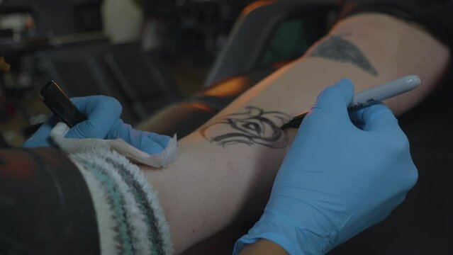 a tattoo artist sketches the pattern for a rose tattoo onto the clients leg in preparation for ink