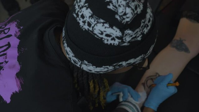 a tattoo artist with dreadlocks sketches a rose pattern onto the leg of his client before applying ink
