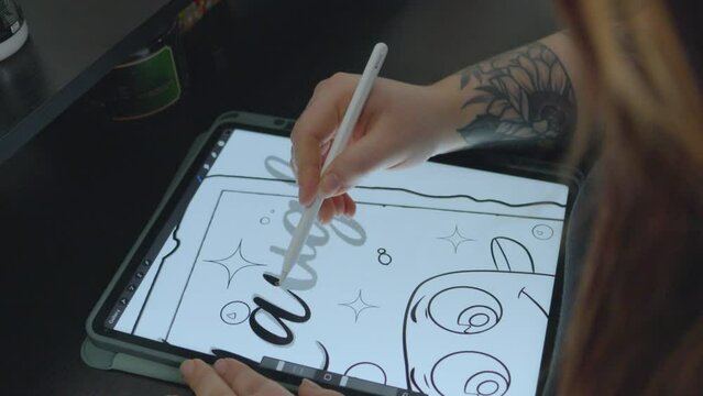 a woman designer with long red hair traces cursive letters on a tablet to prepare the pattern for a tattoo