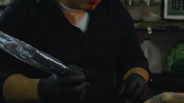 slo motion shot of a tattoo artist wearing black rubber gloves prepares to begin a hand tattoo on a client with other tattoos on her arm