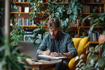 Focused bearded man working on a laptop in a cozy home office surrounded by plants, concept of remote work and creative workspace - obrazy, fototapety, plakaty