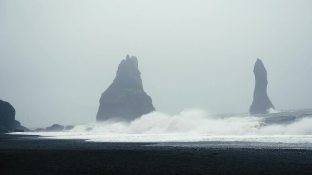Tall dark basalt towers stand out amidst a foggy black sandy beach as waves crash onto the shore. Reynisfjara Iceland.  Shot in glorious slow motion.