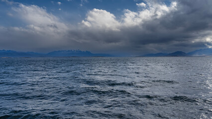 The surface of the blue ocean with small waves and ripples. In the distance, the Andes mountain range is visible against the sky and clouds. The Beagle Channel. Argentina. Tierra del Fuego Archipelago