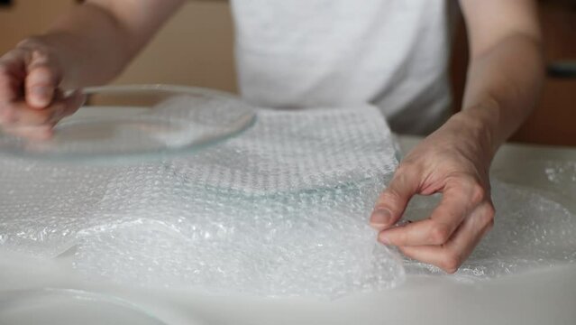 Detail cropped shot of unrecognizable man packing breakable fragile plates glassware in bubble wrap sitting at table preparation to move to new home, cardboard box on background. Concept of new life