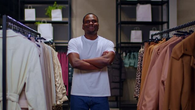 A dark-skinned young man in a white T-shirt stands btw rails with clothes and looks into the frame with his arms crossed, a man smiles with his arms crossed, an affable young man has his arms crossed.