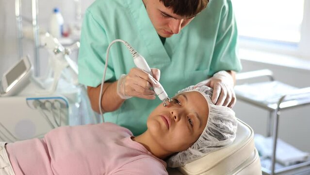 Young male cosmetologist performs hardware facial microcurrent procedure to young female patient