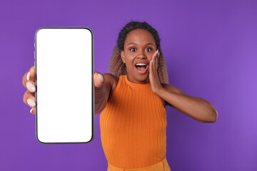 Young attractive shocked African American woman with mobile phone in hand opens mouth after reading unexpected news and experiences wow emotions stands posing in purple studio. White screen