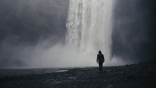 Wide shot of a dark lone hiker walks towards an epic downpour from a water fall amidst dark and moody rocks.  Shot in glorious slow motion. Skógafoss Iceland.