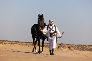 Saudi man in traditional clothing in the desert walking with his black horse by his side
