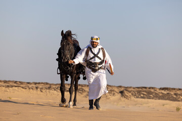 Saudi man in traditional clothing in the desert walking with his black horse by his side