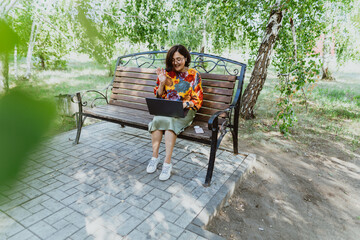 Outdoor office with a cheerful woman conducting video meetings on her computer in nature. Cheerful woman engages in lively video calls on her laptop while sitting on park bench