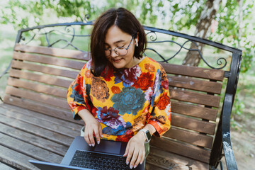 Female professional enjoys remote work on a calm sunny day in a lush green park. Working in fresh air, a woman finds her productive spot on bench amidst the greenery