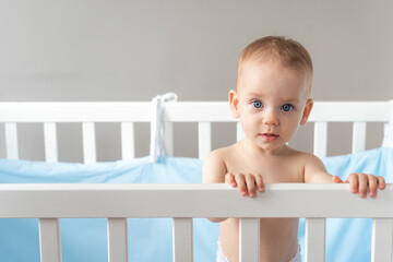 A cute fair baby with expressive blue eyes looks out of a crib