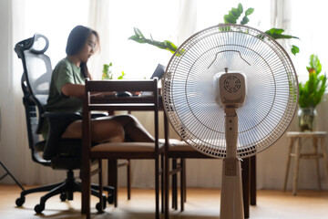 Unhappy Asian woman sitting in front of fan at work suffering from heat in modern house.