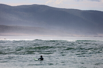 Surfer in the ocean waiting for waves. Beautiful surf conditions in Bicheno, Tasmania. Ocean, surfers and mountains in the background, Australia.