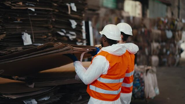 A man with Black skin in a white protective uniform in an orange vest together with his colleague a man with a beard lifts a large rack of cardboard at a waste recycling and sorting plant for waste