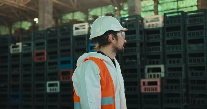 A brunette man with a beard in a protective suit goggles and a helmet in an orange vest walks along black and yellow boxes for sorting and recycling waste at a large factory
